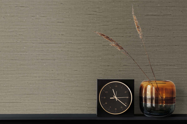 brown grasscloth wallpaper with a gold and black clock next to a bronze vase and two leaves.