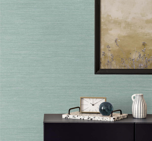 blue sisal wallpaper with a black framed golden poster of wisteria. A black bureau with a sandy and white colored clock, a terrazzo tray with a brown glass paper weight, next to a white and black pinstripe mug.