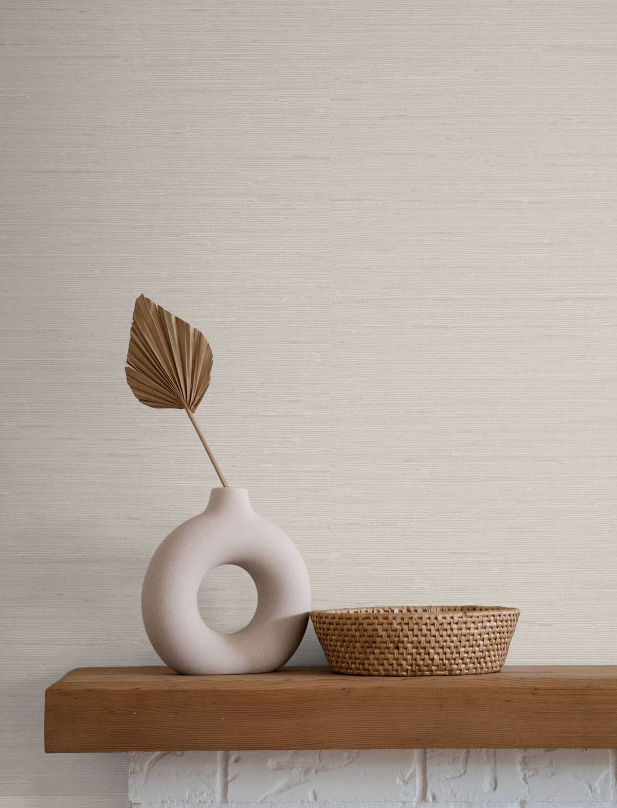 cream grasscloth wallpaper with a white stone vase and brown leaf next to a wicker basket atop a wooden and stone mantel