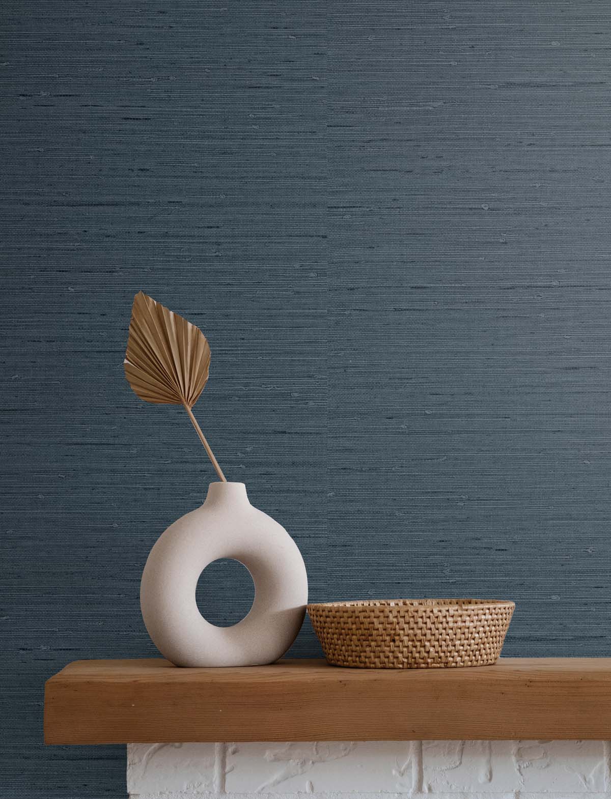 dark blue faux grasscloth wallpaper with a white stone vase, single golden leaf next to an empty wicker basket atop a wooden and white stone fireplace