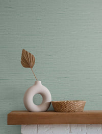 blue green grasscloth wallpaper with a white stone vase and a brown leaf inside, next to a wicker basket atop a wooden and stone mantle