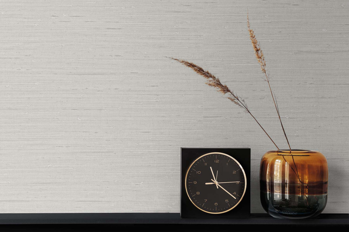 light gray grasscloth wallpaper with a black and gold clock next to a burnished vase with two strands of wheat grass