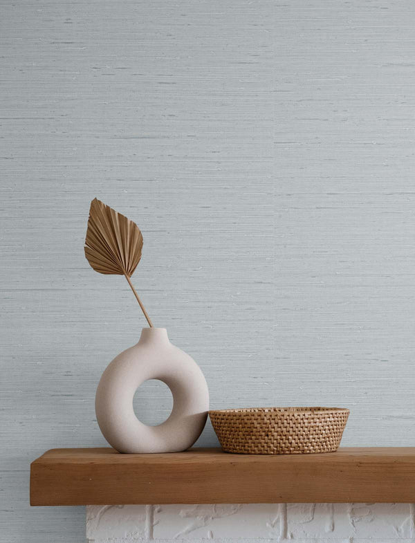 blue gray grasscloth wallpaper with a white stone vase, a brown leaf inside it, next to a wicker basket on top of a wooden and white stone mantle