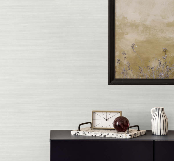 white vinyl wallpaper made of embossed grasscloth with a black framed golden poster of wisteria. A black bureau with a sandy and white colored clock, a terrazzo tray with a brown glass paper weight, next to a white and black pinstripe mug.