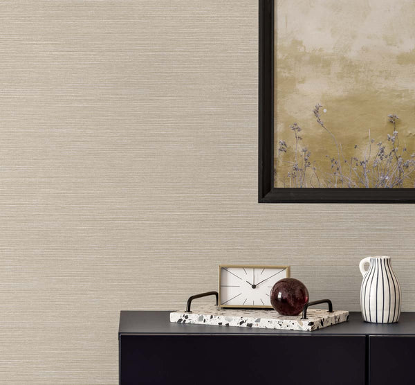 woven beige faux grasscloth wallpaper with a black framed golden poster of wisteria. A black bureau with a sandy and white colored clock, a terrazzo tray with a brown glass paper weight, next to a white and black pinstripe mug.