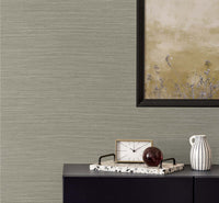 taupe grasscloth wallpaper with a black framed golden poster of wisteria. A black bureau with a sandy and white colored clock, a terrazzo tray with a brown glass paper weight, next to a white and black pinstripe mug.