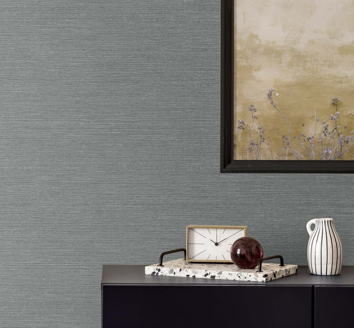 gray grasscloth wallpaper with a black framed golden poster of wisteria. A black bureau with a sandy and white colored clock, a terrazzo tray with a brown glass paper weight, next to a white and black pinstripe mug.