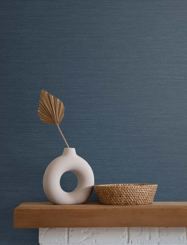 navy blue grasscloth wallpaper with a white stone vase holding a golden brown leaf, next to a beige wicker basket, atop a wooden mantle and white brick fireplace.