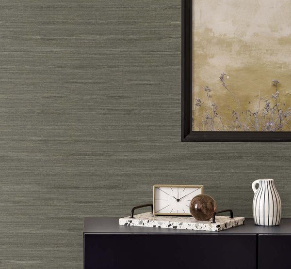 dark brown grasscloth wallpaper with a black framed golden poster of wisteria. A black bureau with a sandy and white colored clock, a terrazzo tray with a brown glass paper weight, next to a white and black pinstripe mug.