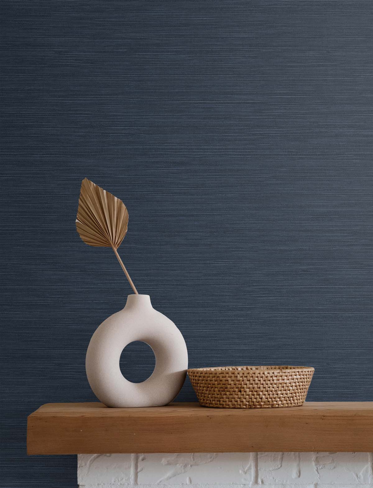 dark blue wallpaper with a white stone vase holding a golden brown leaf, next to a beige wicker basket, atop a wooden mantle and white brick fireplace.