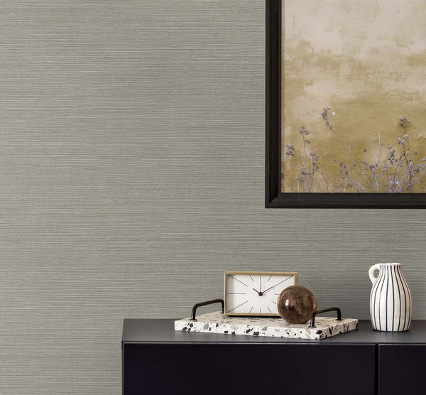 plain gray grasscloth wallpaper with a black framed golden poster of wisteria. A black bureau with a sandy and white colored clock, a terrazzo tray with a brown glass paper weight, next to a white and black pinstripe mug.