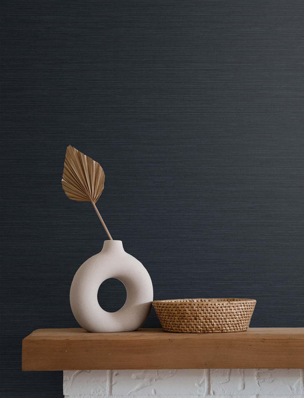Dark blue grasscloth wallpaper with a white stone vase holding a golden brown leaf, next to a beige wicker basket, atop a wooden mantle and white brick fireplace.