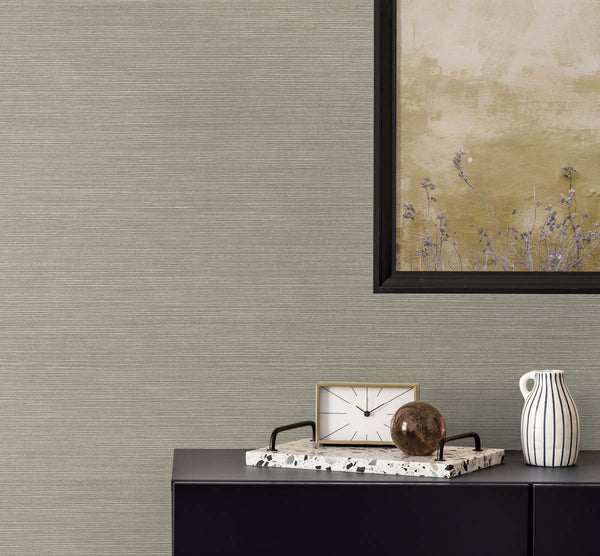 plain beige grasscloth wallpaper with a black framed golden poster of wisteria. A black bureau with a sandy and white colored clock, a terrazzo tray with a brown glass paper weight, next to a white and black pinstripe mug.